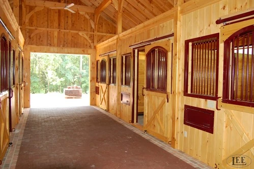 Bright barn aisle featuring light wood construction, an arched truss ceiling, and a dark brick floor. The stalls have dark red metal sliding doors with barred tops and cross-buck wood bottoms, leading to a large open doorway at the far end.