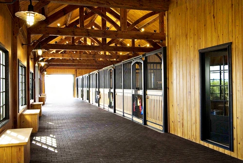 Elegant timber-frame barn aisle with black metal stall fronts, mahogany lower panels and bright daylight at end