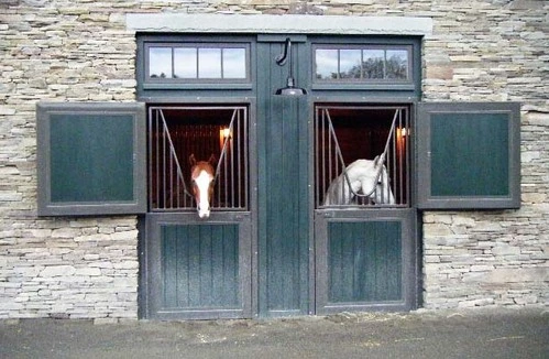 Stacked stone wall barn with dark teal sliding doors and two white horses visible inside stalls