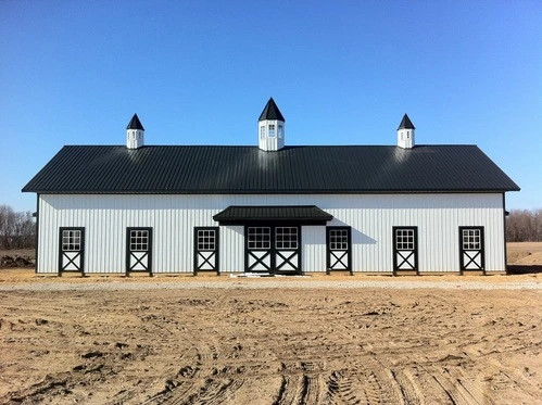 White metal barn with black roof and three cupolas featuring X-pattern stall doors along front