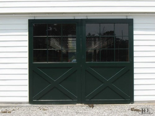 Dark green double doors with X-pattern and upper glass panels on white barn siding