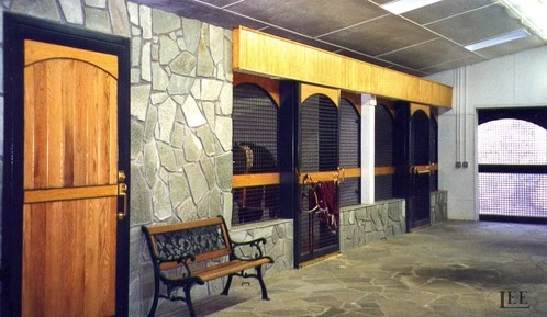 Natural wood stall fronts with arched tops and wire mesh panels along stone accent wall barn aisle