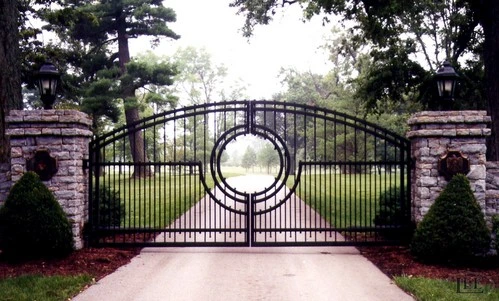 Black ornamental double gates featuring large circular center medallion between brick pillars with landscaping