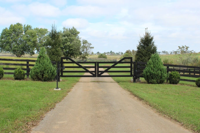 Dark wood farm-style gate spanning dirt driveway between black board fencing with evergreen trees