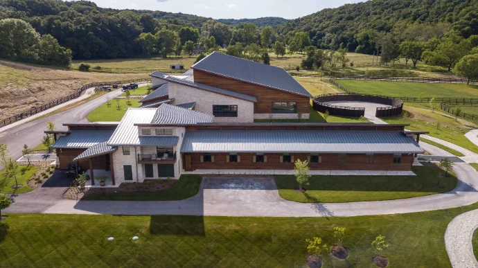 Aerial view of modern barn complex with white metal roofs, stone accents and circular driveway in rural landscape