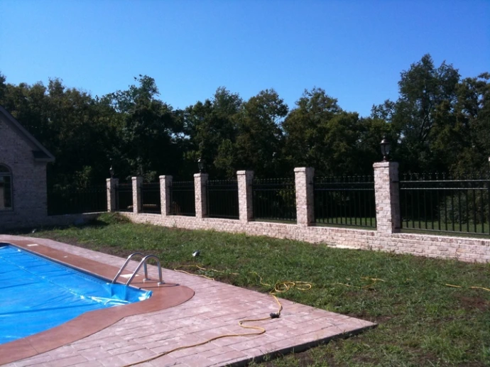 Black metal vertical bar pool fence along curved edge of in-ground pool with white stone pillars and green lawn