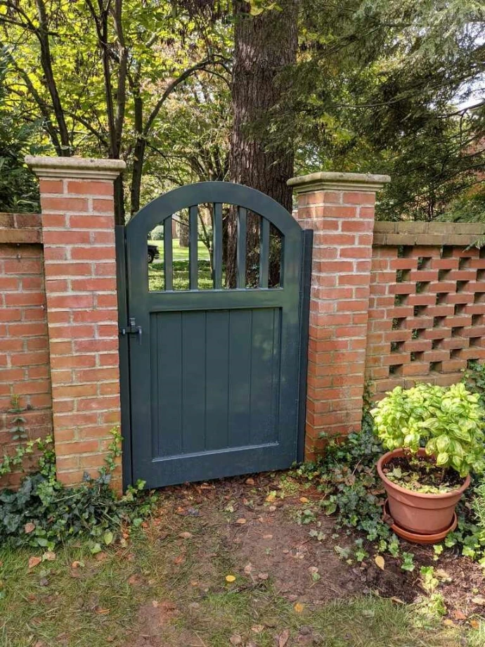 Dark blue-gray arched garden gate with vertical bars and solid lower panel set between red brick pillars with hedge