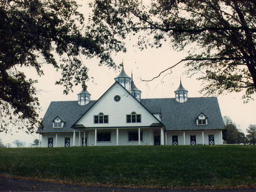 White barn with dark roof, multiple cupolas, dormers and symmetrical stall openings on green lawn