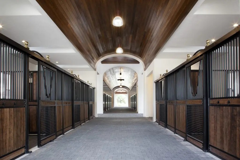 Luxury Barn Interior - Hero Image: Long elegant barn aisle with black metal stall fronts, rich mahogany lower panels, white walls, dramatic wood plank ceiling with pendant lights, gray paver flooring and arched opening at end