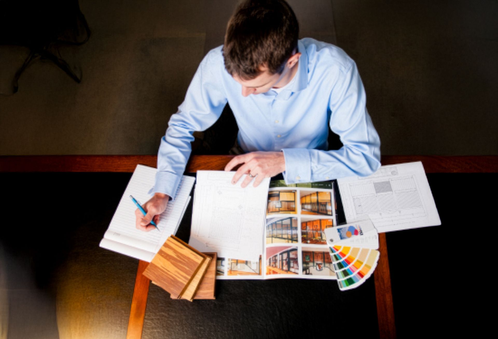 How to Get Started - Step 3 (Review with experts): Aerial view of person in blue shirt reviewing floor plans and material samples at desk with barn photos