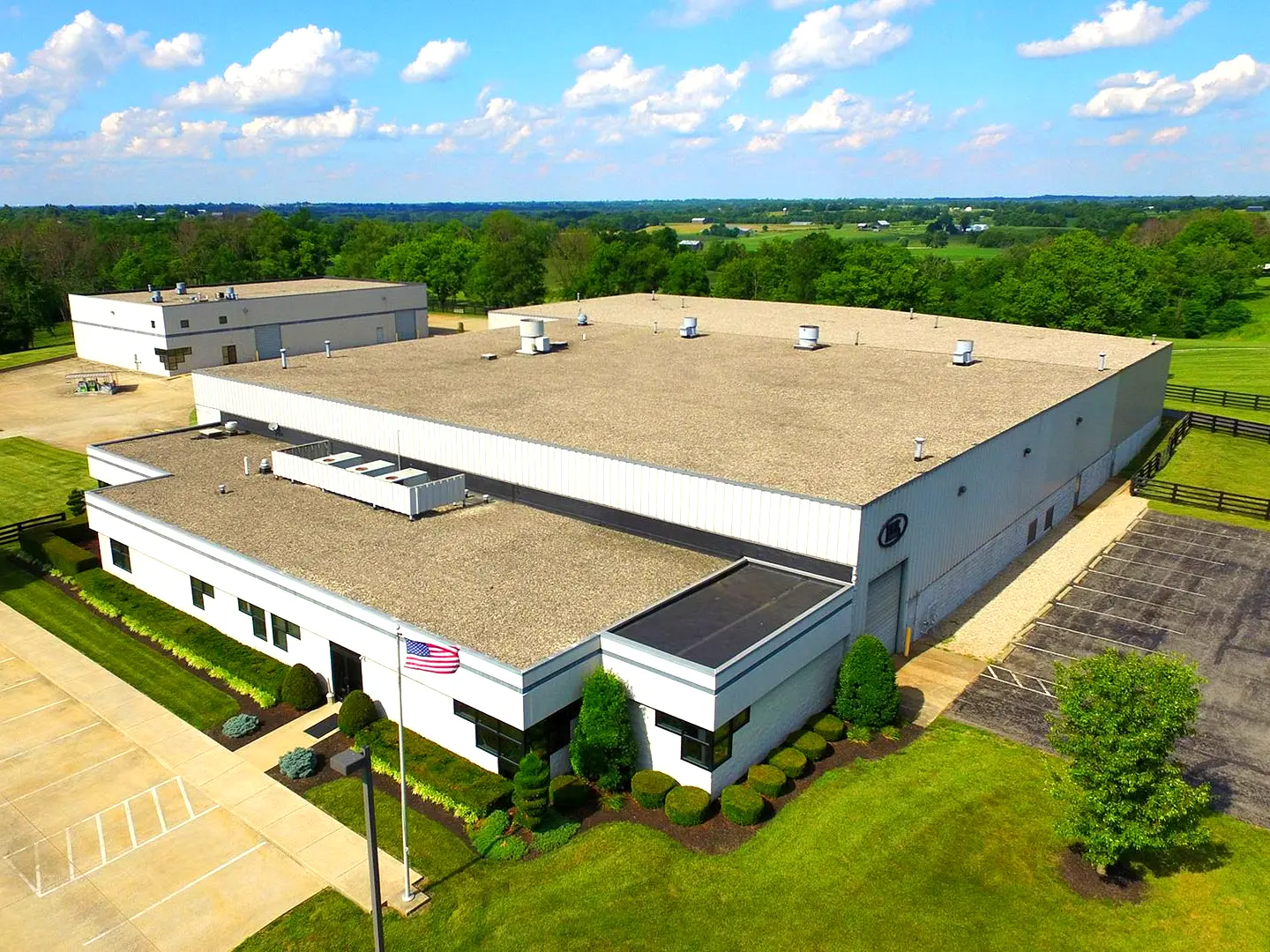 Manufacturing Facility: Aerial view of large white and gray industrial facility with flat roof, American flag at entrance and surrounding green landscape with horse fencing