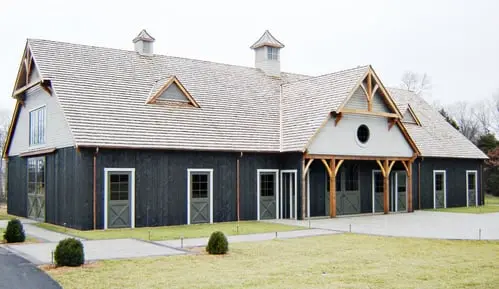 Why Section Image: Modern barn with dark vertical siding, white trim, two cupolas and multiple white-framed door openings along front facade on green lawn