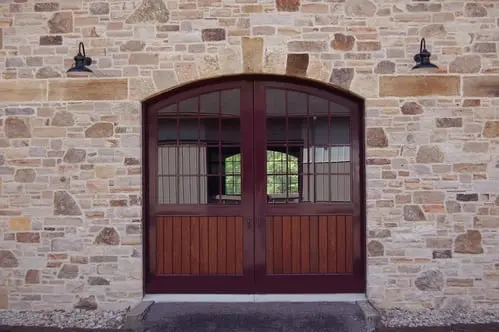 How Section Image: Close-up of arched mahogany double doors with multi-pane windows set in tan stone wall with black wall-mounted lanterns